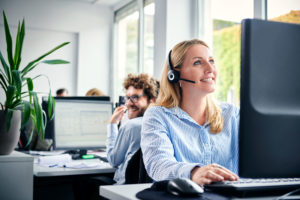 Aufnahme Kundenservice. Helle freundliche Aufnahme in einem Büro mit großen Fenstern. Eine Frau sitzt mit einem Headset an einem Bildschirm an einem Schreibtisch und lächelt. Im Hintergrund sitzt ein weiterer Mitarbeiter an einem Schreibtisch und guckt lächelnd über die Schulter zur Kamera.