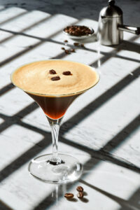 Ein Kaffe als Cocktail in einem Martini Glas garniert mit Kaffeebohnen auf weißem Marmor in einem schrägen Licht durch Jalousinen.