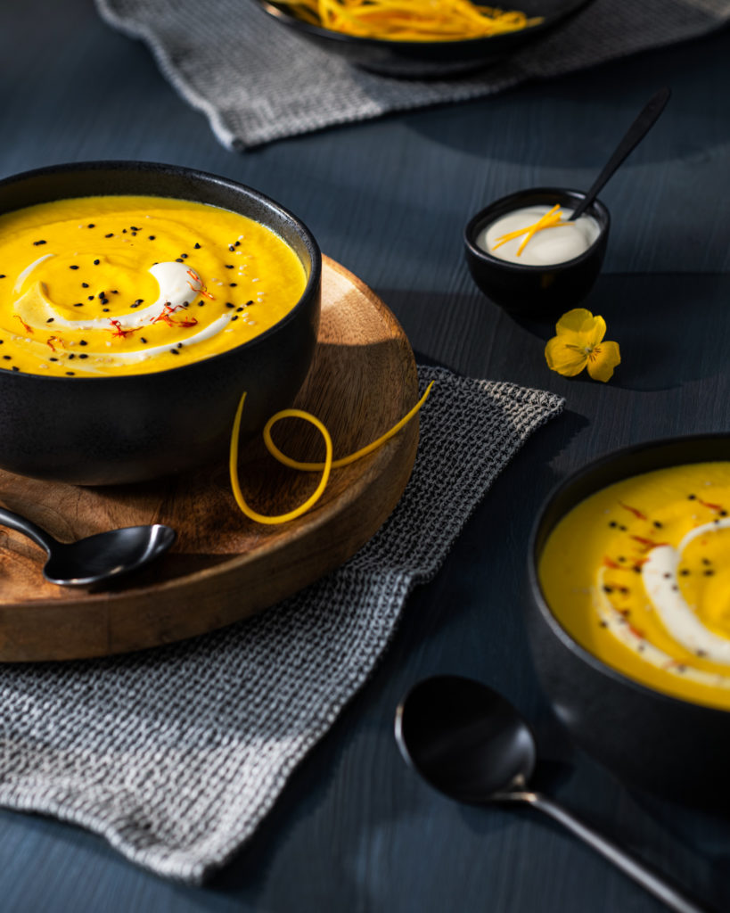 Eine gelbe Cremesuppe in schwarzen Schalen auf dunkelblaum Holz.