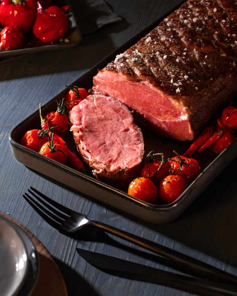 Ein Braten in einer Backform mit ganzen Tomaten auf einer Holzpolatte im steilen Licht.