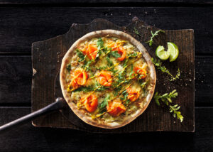 Eine Lachs-Rucola-Pizza auf einem dunklen Holzbrett aus einer organischen Baumscheibe mit großem Pizzaschieber aus Metall.