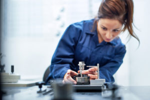 Industrieaufnahme. Eine Frau in einem Blaumann hält in den Händen ein Bauteil. Der Fokus der Kamera liegt auf dem Bauteil auf dem Tisch. Die Umgebung ist hell und modern.